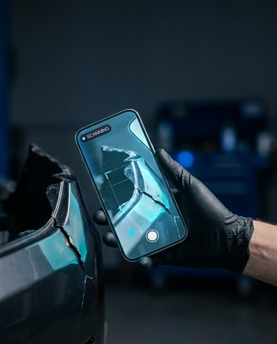 Close-up of a gloved hand holding a phone beside a torn bumper corner, the damage framed on screen by the scanner